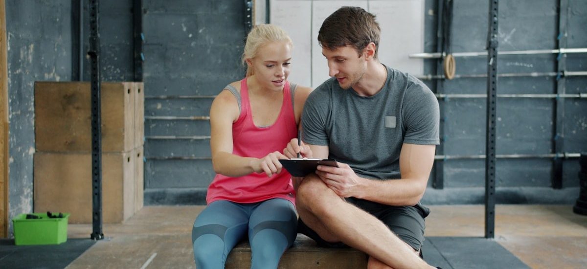 Man professional instructor is talking to young woman and writing in chart relaxing after training, people are waring trendy sports clothing and footwear.